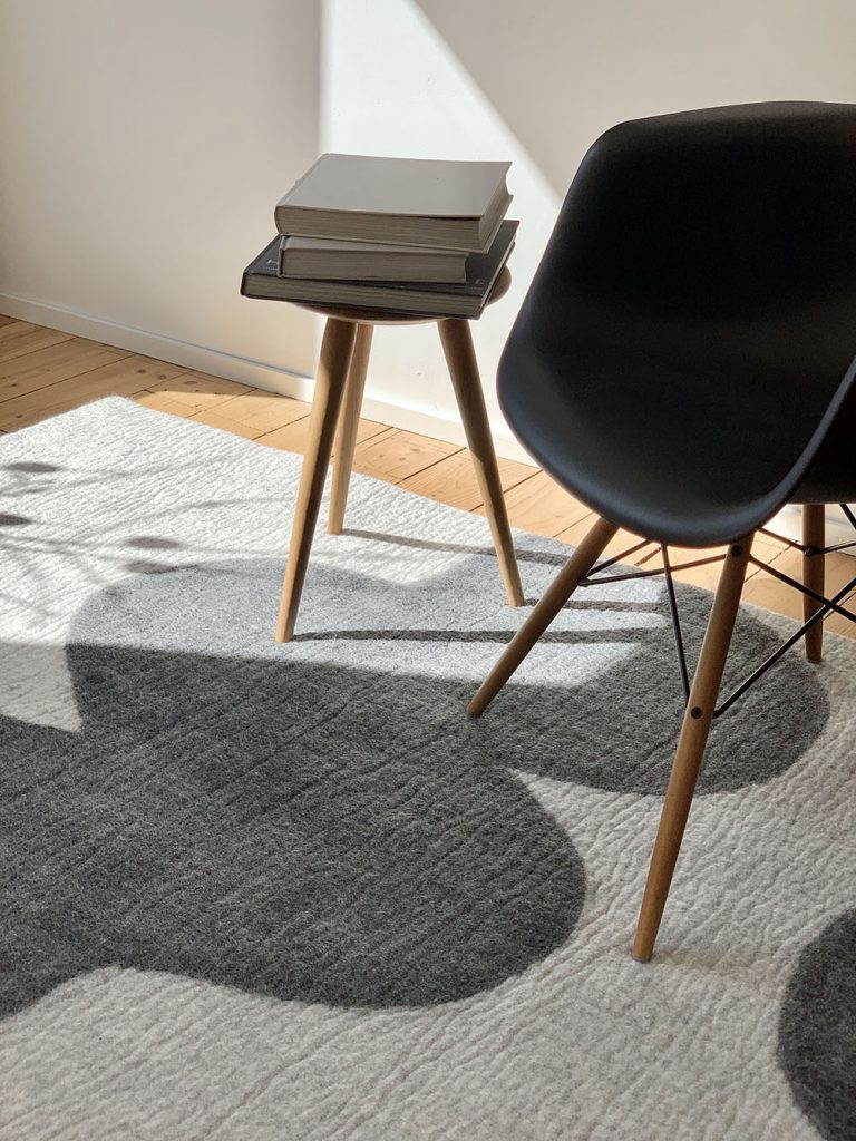 A modern, minimalist interior featuring a felt rug with abstract black and grey shapes. A black Eames arm chair with wooden legs is placed next to a small wooden side table, which holds a stack of books. Sunlight streams in from a nearby window, casting soft shadows across the floor, enhancing the texture of the rug. The space has a clean, contemporary feel with natural light and simple, functional furniture.